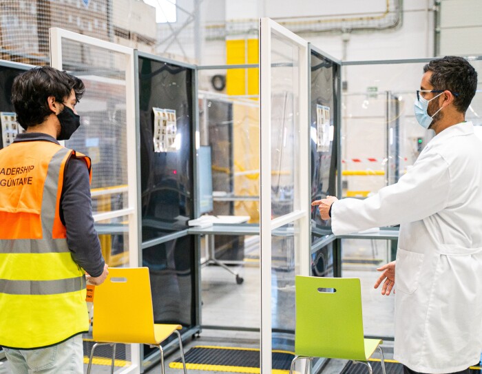 Gianluca Manuli dando indicaciones a un asociado antes de hacerse la PCR. Hay tres puestos cubiertos con mamparas de plástico. El doctor Manuli va con una bata de médico blanca y lleva mascarilla y gafas negras. Da instrucciones a un asociado que va con un chaleco amarillo y naranja, tejanos y mascarilla negra.