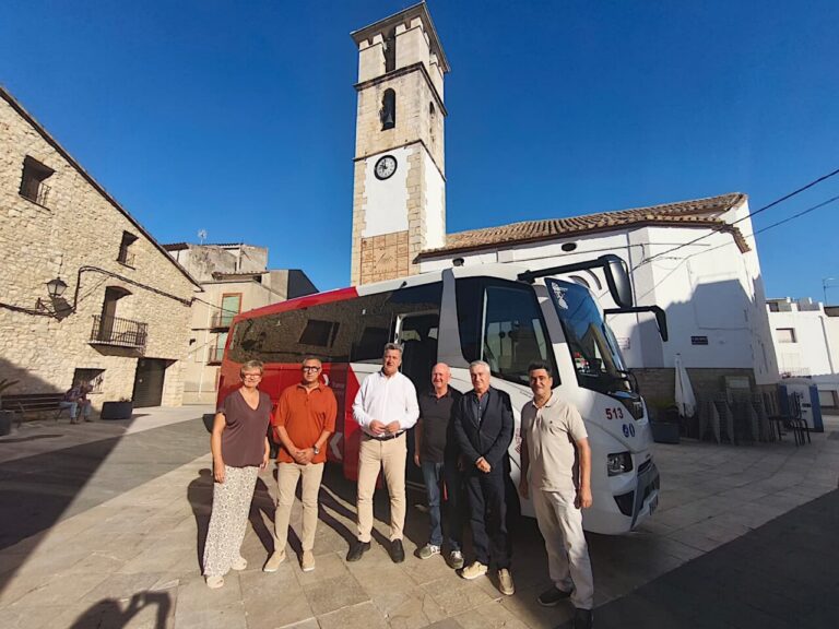 Autobús estacionado junto a varias personas, frente a una torre con reloj en Tírig.