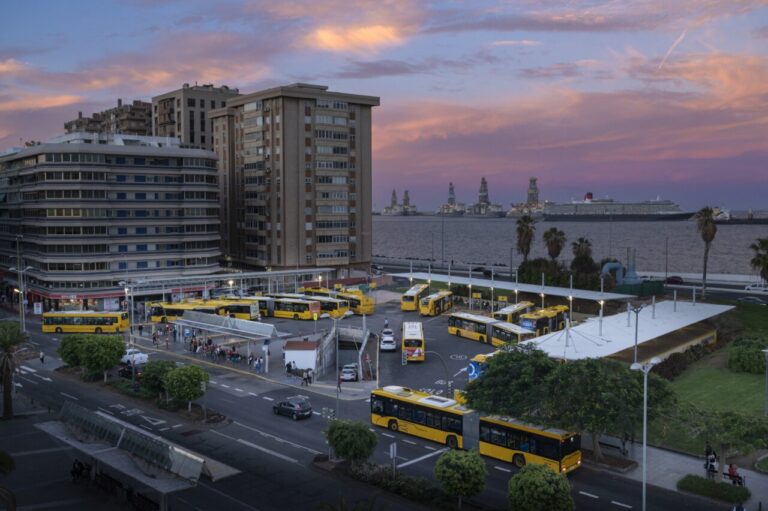 Autobuses amarillos estacionados en una terminal urbana junto al puerto al atardecer.